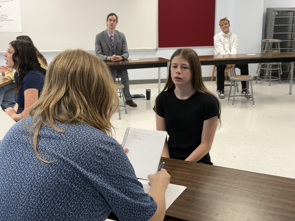 A classroom with several students. One student is seated while another stands and reads aloud from a paper.