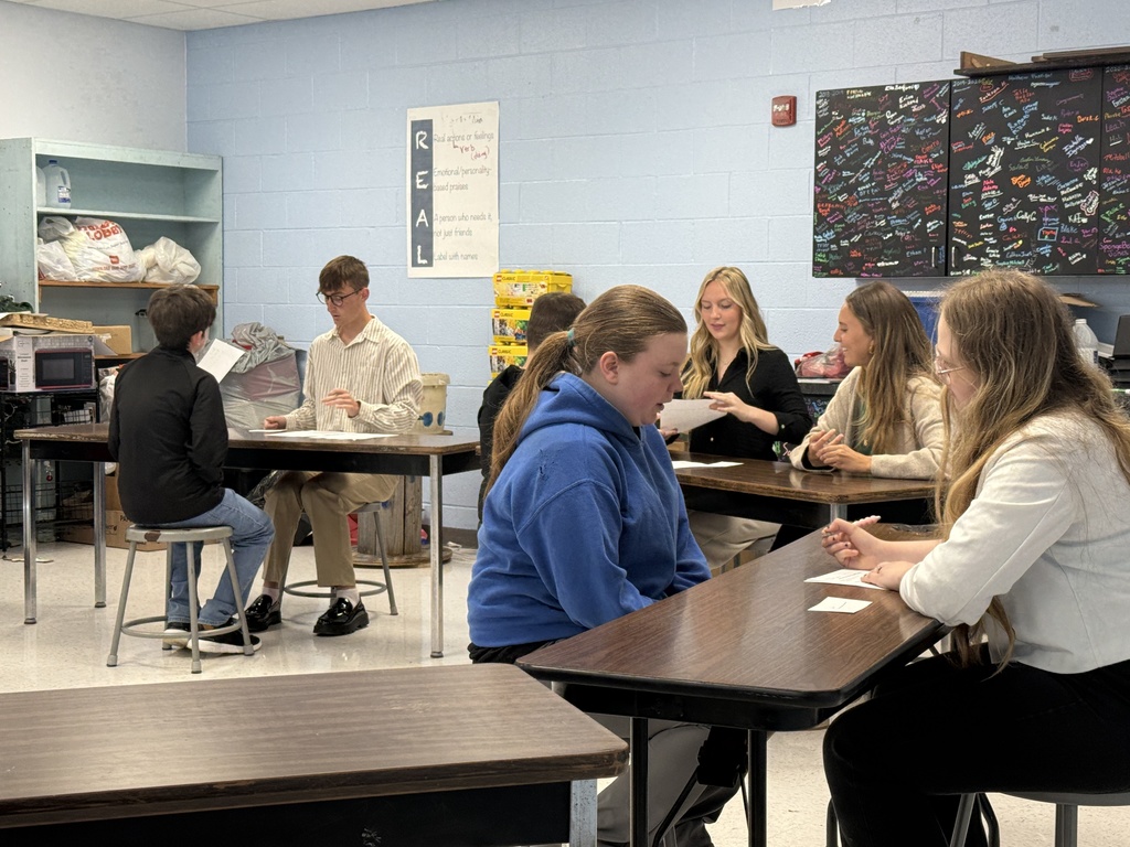 Several students in a classroom, seated around tables. One student is speaking, while others are listening. Blue walls, shelves, and tables.