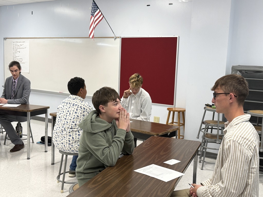 Five individuals in a classroom. Four are seated around tables; one stands by a whiteboard.
