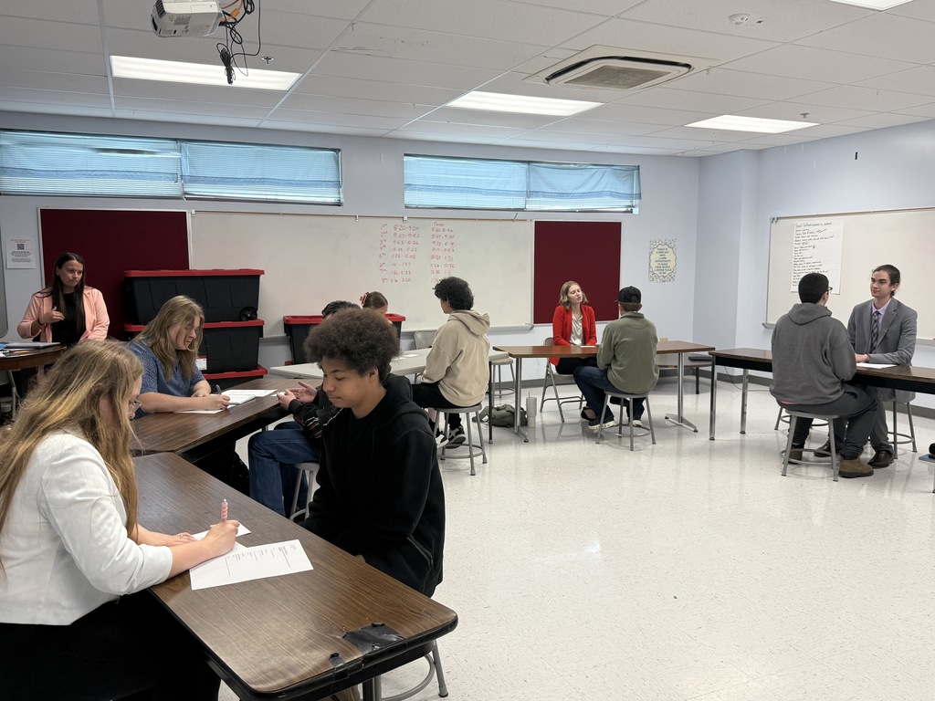A classroom with tables, chairs, whiteboards, and students. Some students are seated while others stand, and one student writes on paper.
