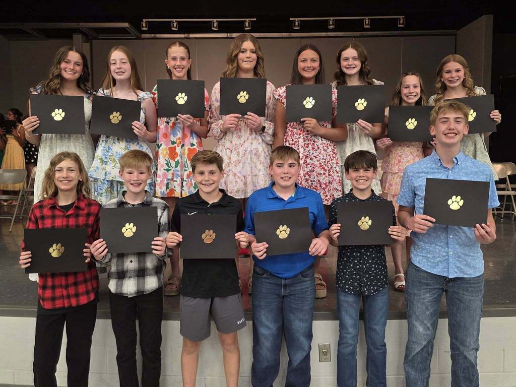 Group of children holding black and yellow certificates on stage. Some wear casual clothing.