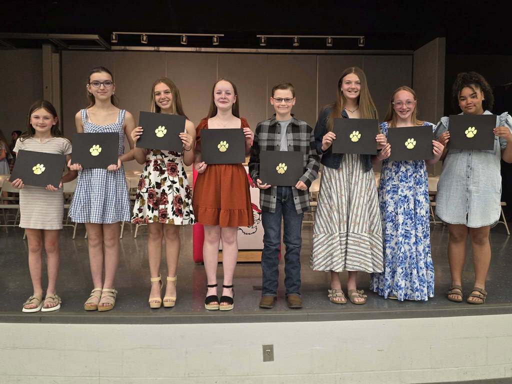 A group of students stands on stage, holding certificates. They wear various outfits, including dresses, sandals, and sneakers. The stage has a black backdrop.