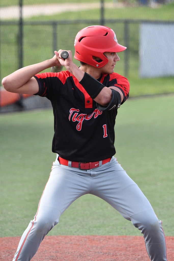 A baseball player in a red helmet and black uniform with "Tigers" text swings a bat on a baseball field.