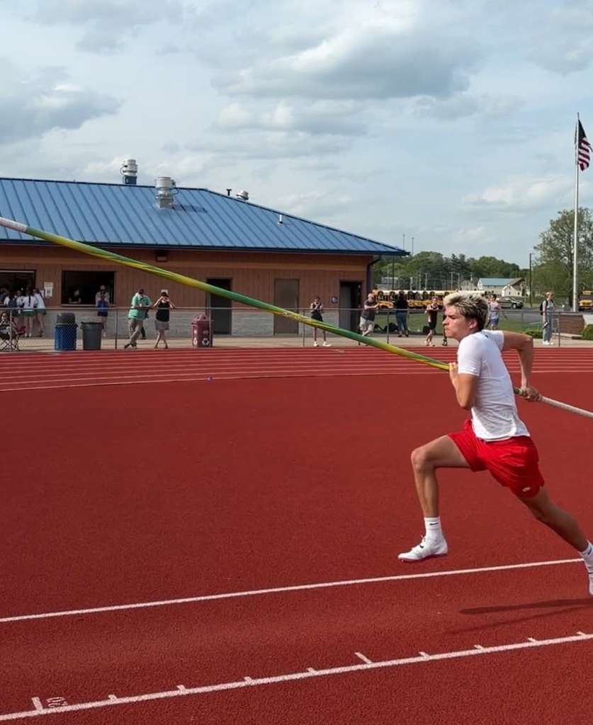 A man runs on a red track, holding a long pole for a javelin throw. Behind him, a building and people are visible.