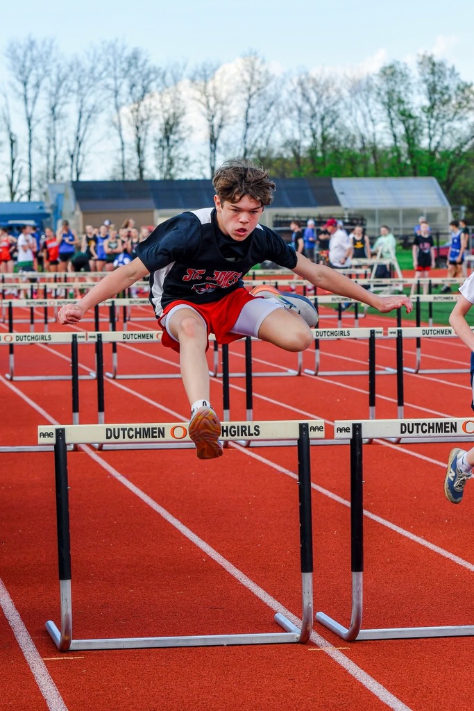 A boy jumps hurdles on a track. Spectators in the background watch. Trees line the edges of the track.