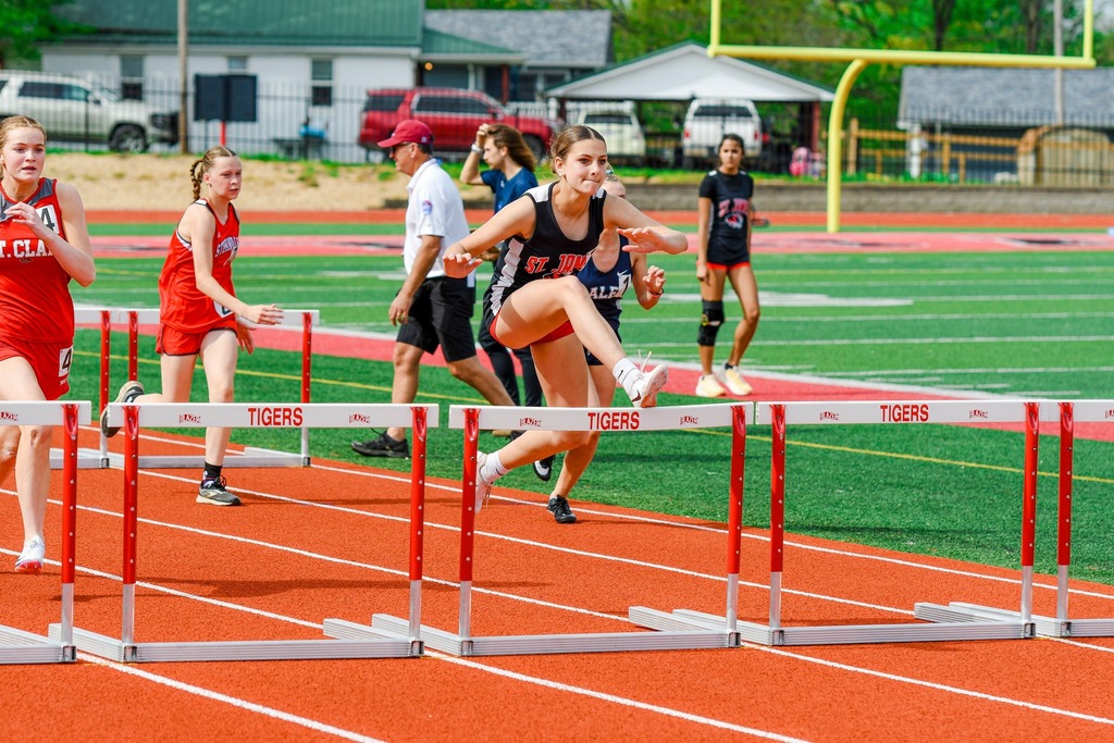 A group of athletes compete in a hurdling event on a track, with hurdles and a grassy field.