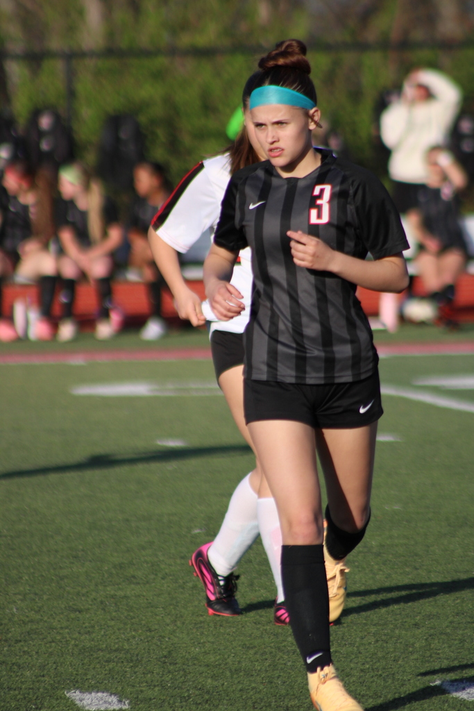 Two female soccer players, one wearing a black and white striped jersey, running on a field. In the background, spectators are seated.