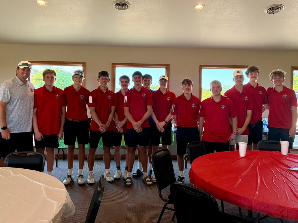 A group of athletes in red shirts and black shorts pose indoors, with a coach in a white shirt. Behind them are chairs and tables, and two large windows.