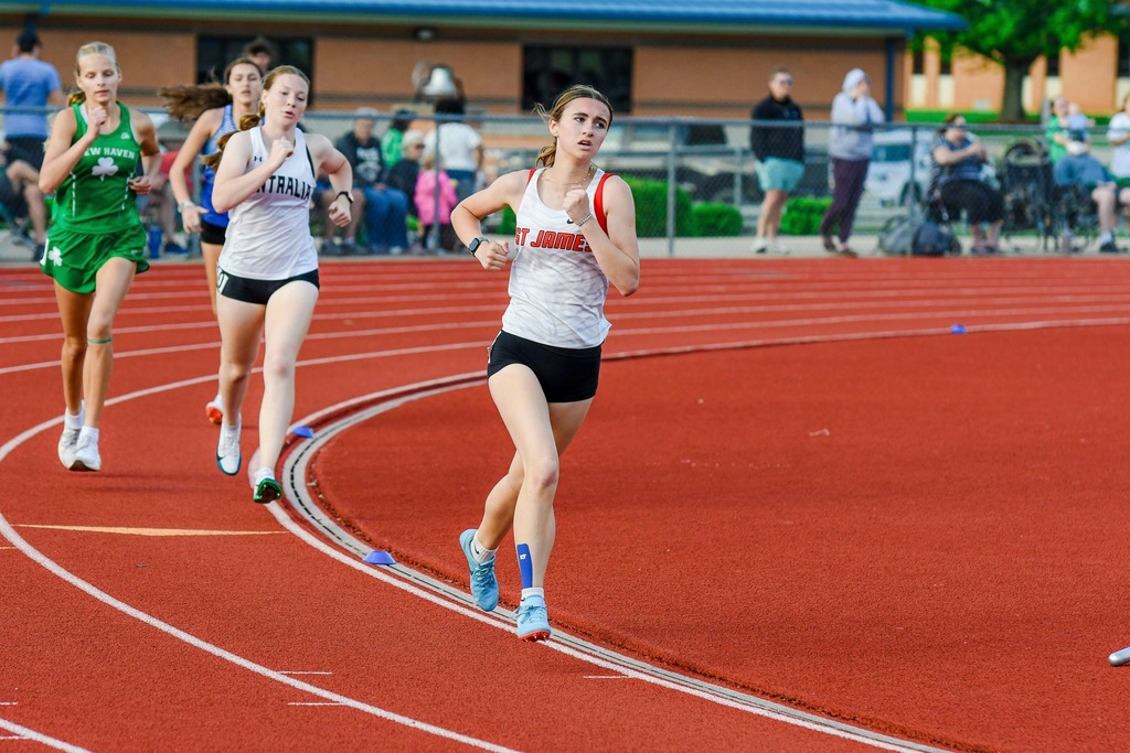 Women running on a track; two in white shirts, one in green, all in sneakers. Audience blurred in background.