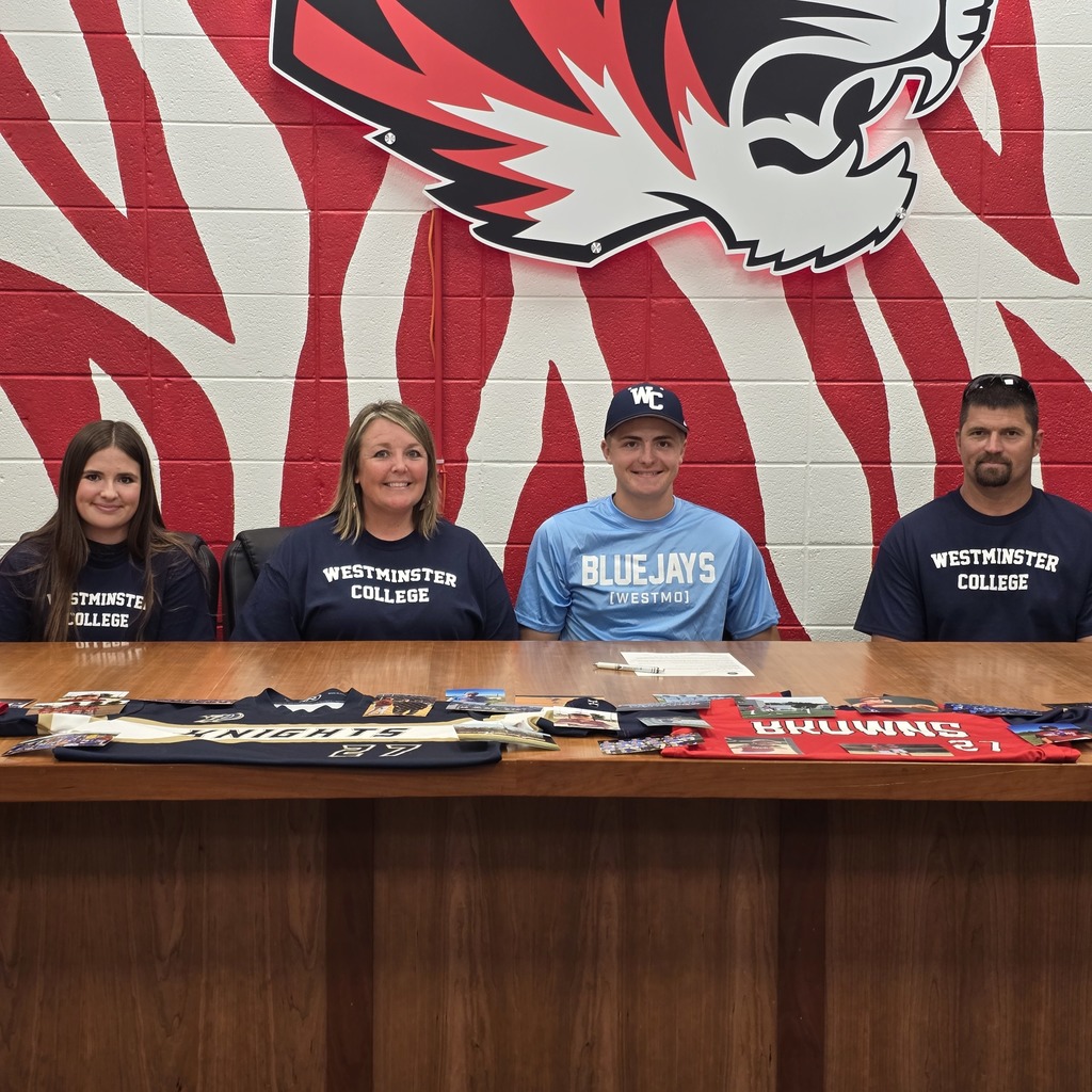Four individuals sit at a table with a lion emblem on a striped background. Each wears a "Westminster College" shirt.