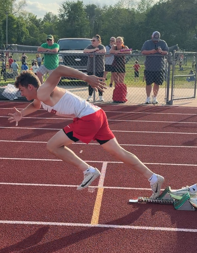 An athlete jumps on a red track, in a starting position. People are standing behind a fence.