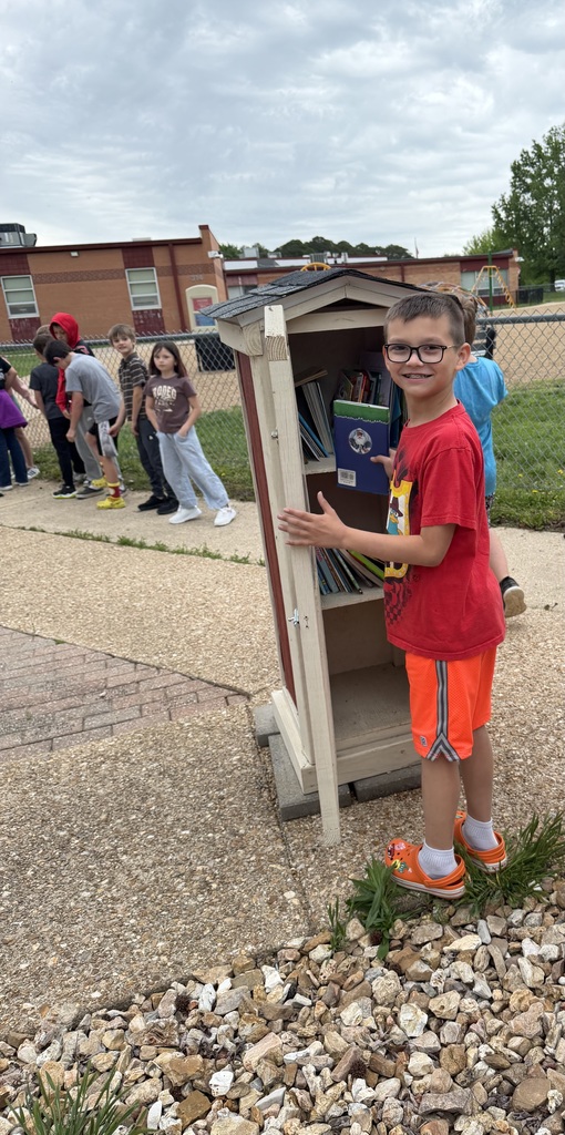 A young boy in glasses stands next to a small bookshelf outdoors. Other children stand behind a fence.