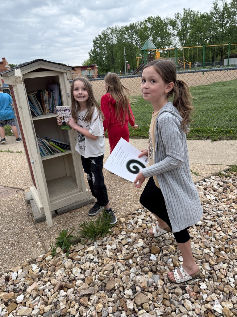 A girl with ponytails holds a book and paper with a symbol. Another girl holds books. Small shelf filled with books behind them. Background: grassy area with a fence.