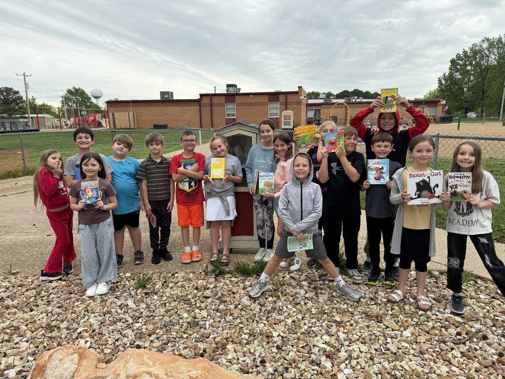 Students pose holding books in front of playground and school.