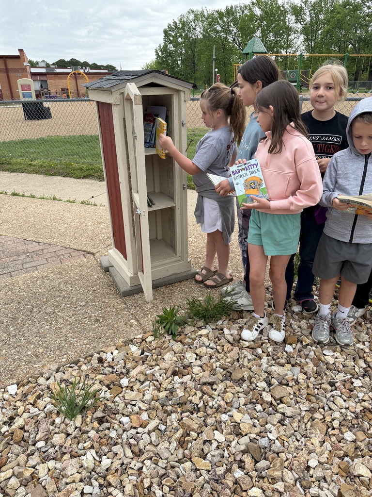 Six children are looking at a small, red and white structure. One child holds a book while reaching into it.