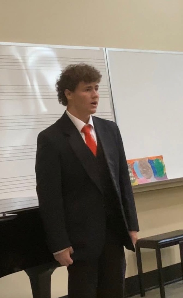 A man in a black suit and red tie stands next to a piano and a whiteboard.