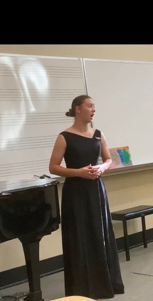 A woman in a black dress stands next to a piano in a room with a white backdrop.