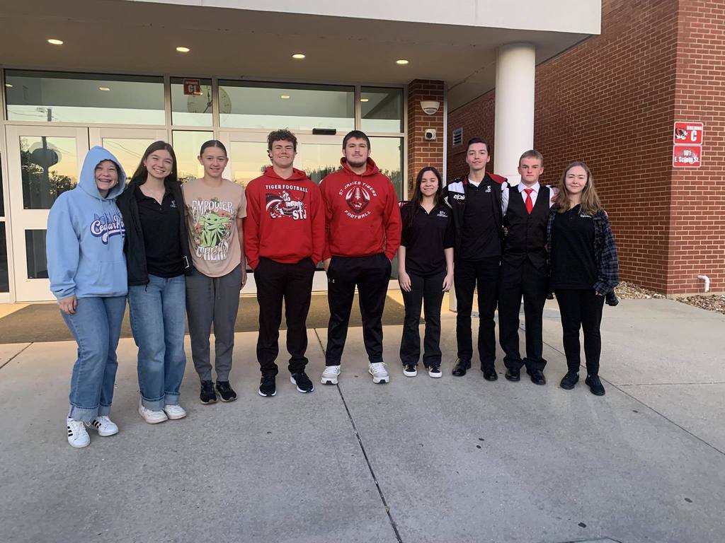 Eight people stand outside a building, smiling. Four wear red hoodies, two in dark jackets, one in a blue hoodie, and one in a white shirt.