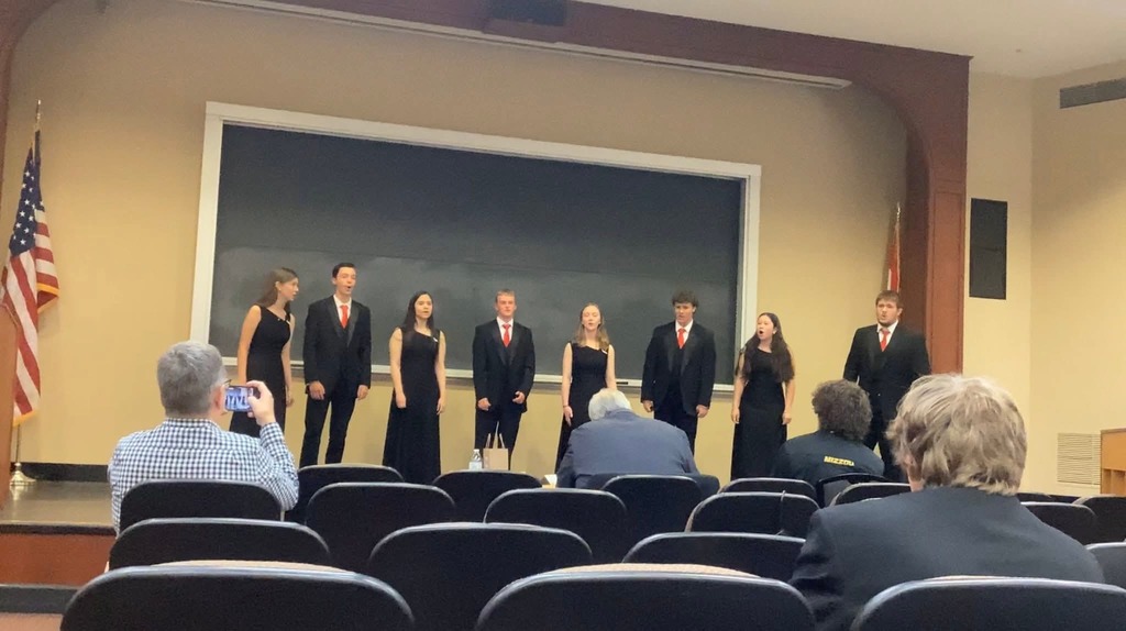 Seven people in suits and dresses stand on stage with an audience seated in chairs in front.