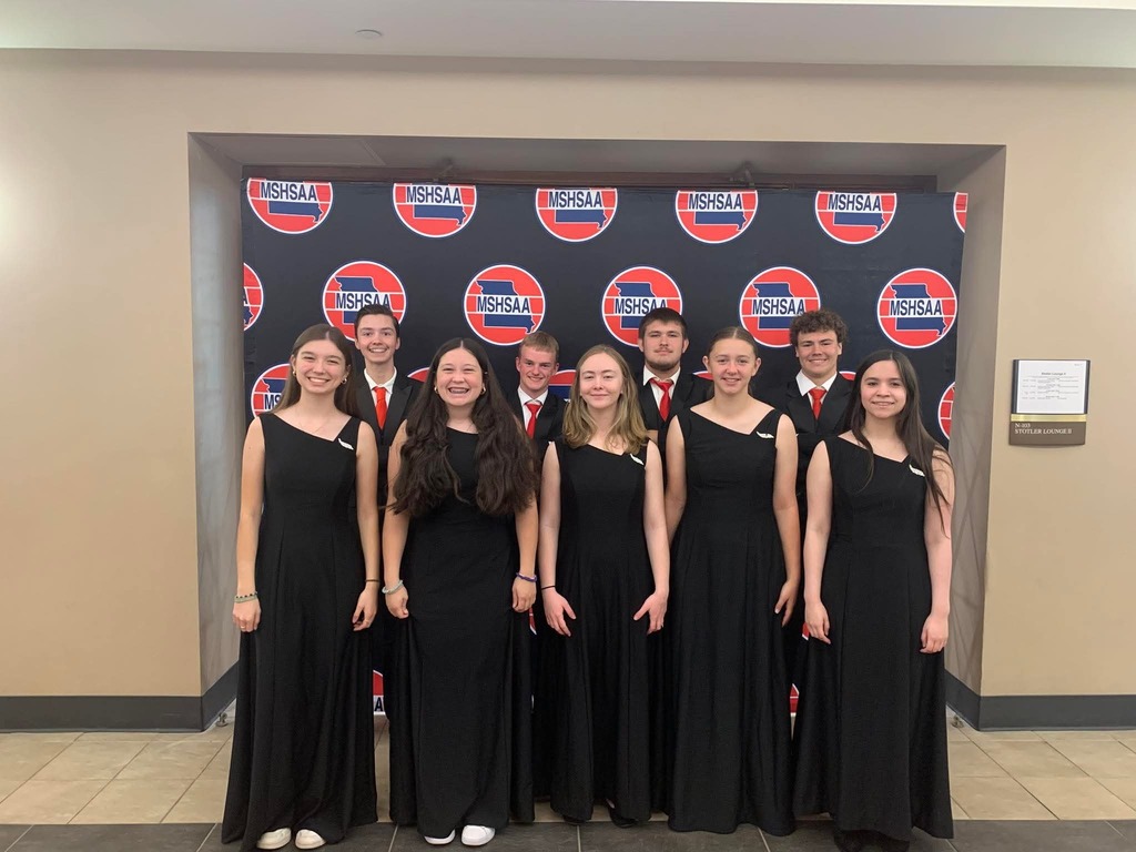 A group of students in black dresses standing in front of a wall with red and white circular designs.