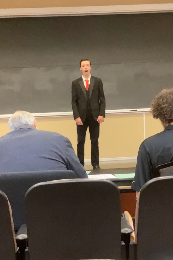 A man in a suit and tie stands on a stage, singing. Behind him is a blackboard, and in front, two people are seated.