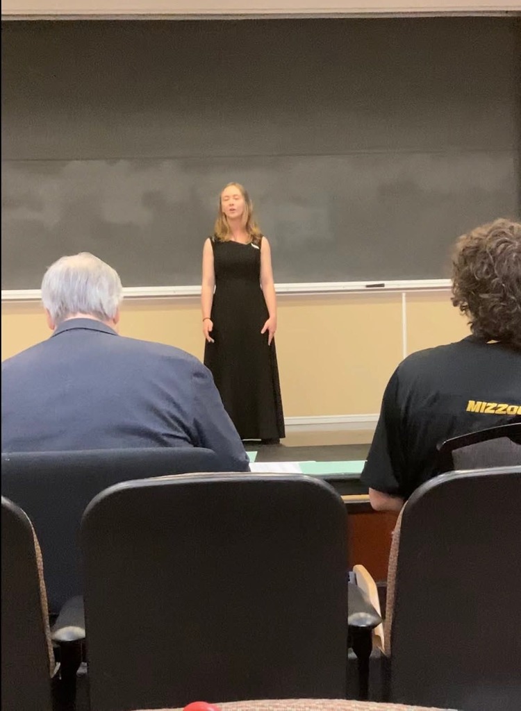 Woman in black dress stands on stage before a blackboard. Two people sit in chairs, one in a blue shirt and the other in a black MIZZOU shirt.