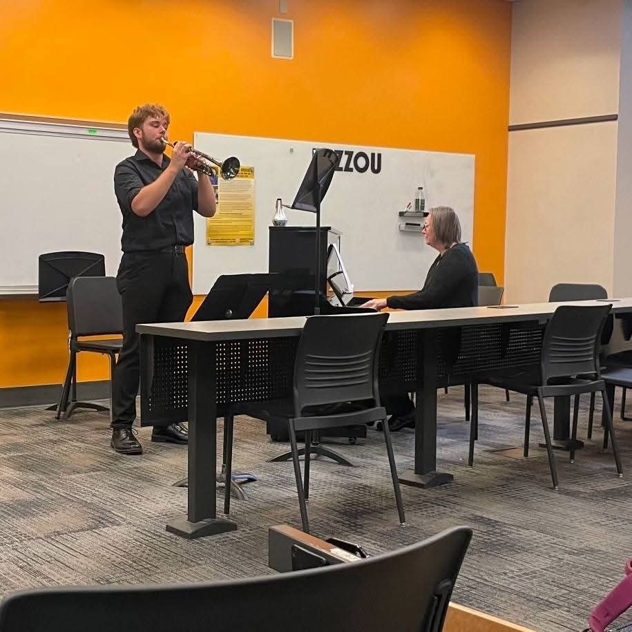 A musician plays a trumpet in a room with chairs. A woman is seated at a desk.