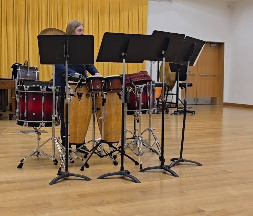 A person plays percussion instruments, including drums and gongs, in a room with a wooden floor and yellow curtains.