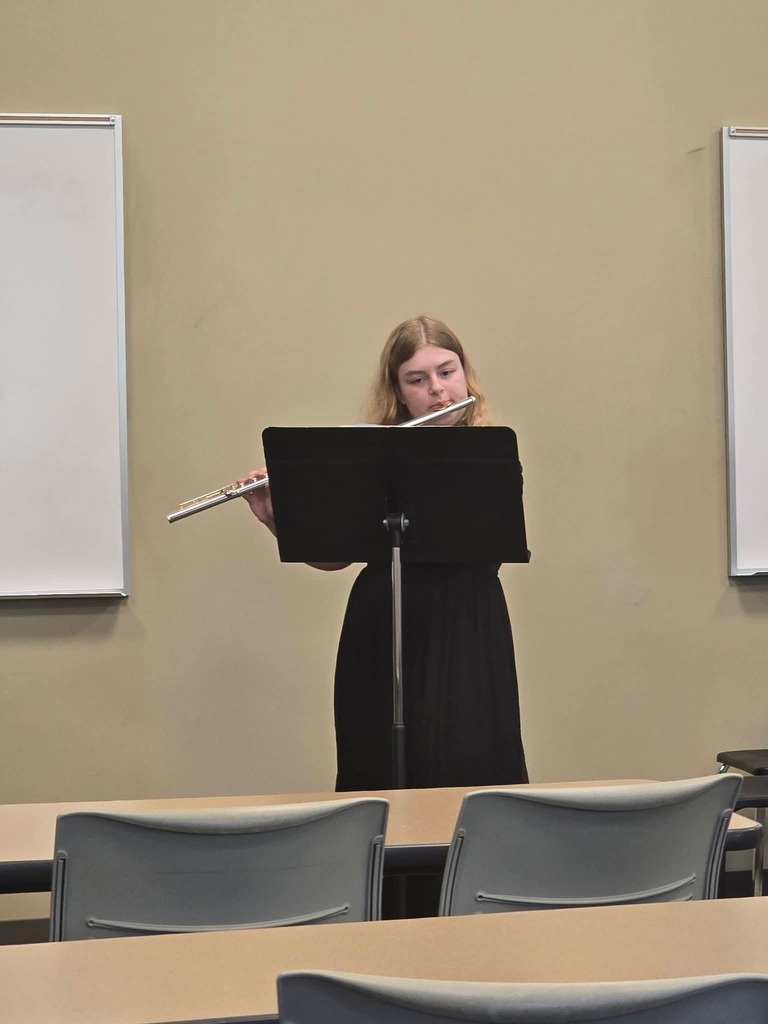 A woman in a black dress plays the flute in a classroom, with chairs and whiteboards in the background.