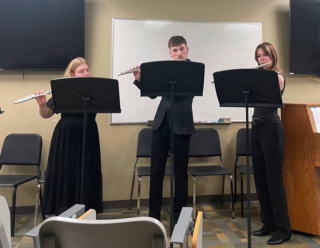 Three young adults, two women and one man, play flutes at music stands in a classroom.