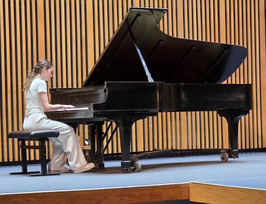 A person sits on a piano bench and plays a black grand piano on a stage with a wooden backdrop.