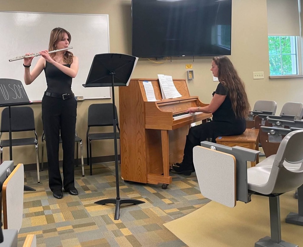 A woman standing holding a flute in a room with a piano. A second woman plays the piano.