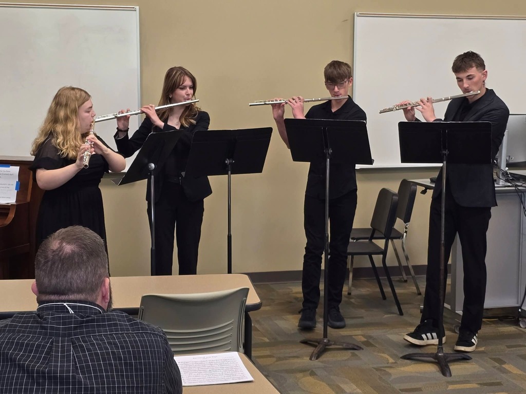 Four musicians play flutes in a classroom with a teacher. The room has tables, chairs, and whiteboards.