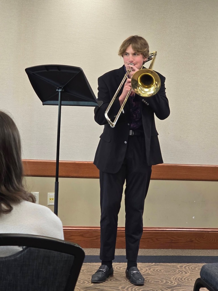 Person in a suit playing trombone in front of a music stand. An audience member watches.