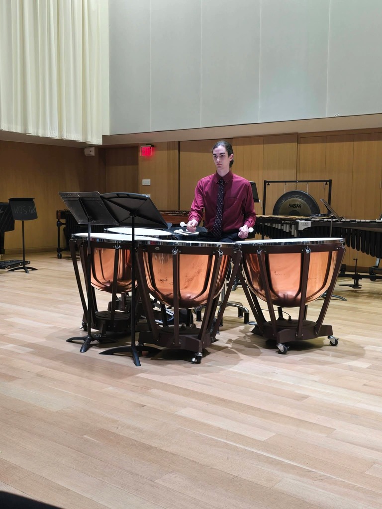 A musician plays a pair of timpani in a music room with a wooden floor.