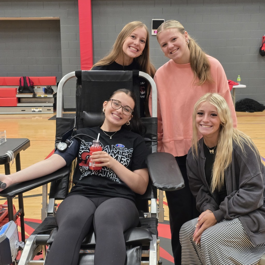 Four women, one in a wheelchair, smile at the camera in a gymnasium with red bleachers.
