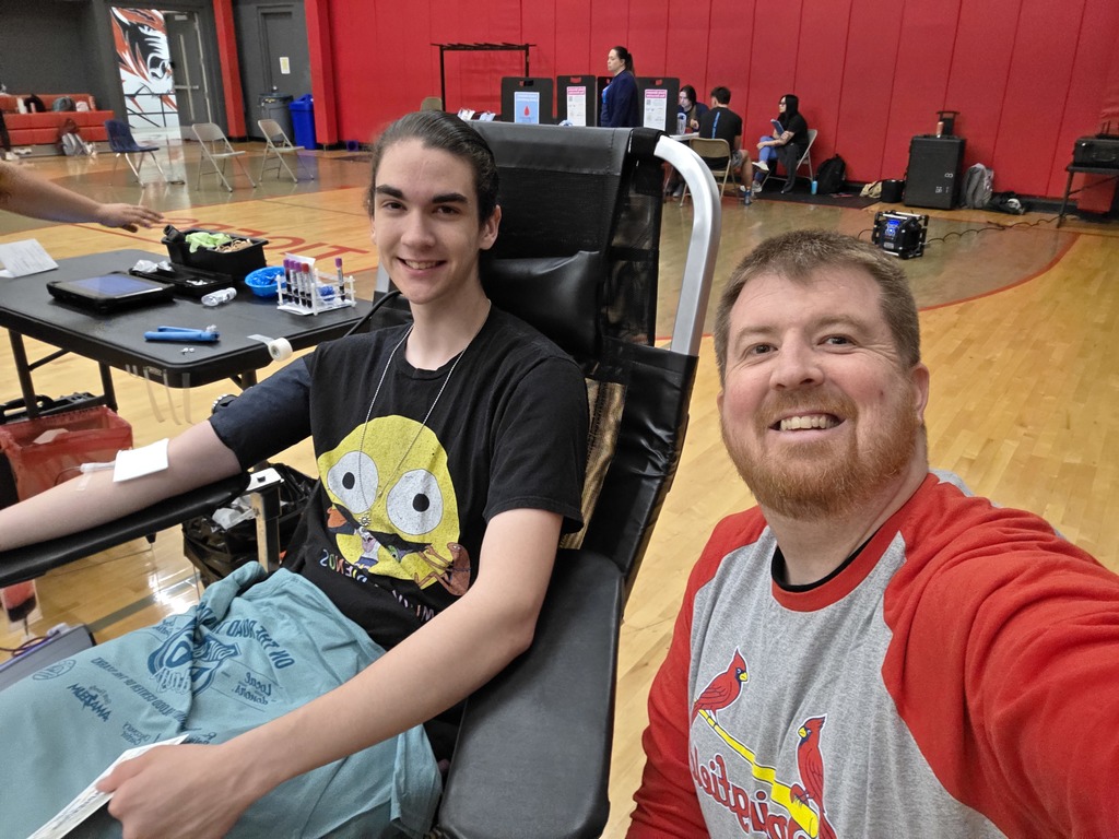 Two males sit in chairs in a gym. One is in a black shirt and is giving blood. THe other is in a red shirt. 