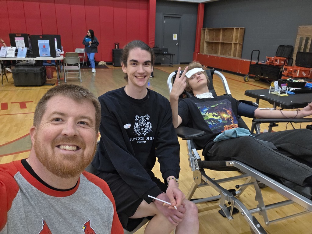 Three people in a room. One man is sitting in a chair, another is standing, and the third is lying on a bed.