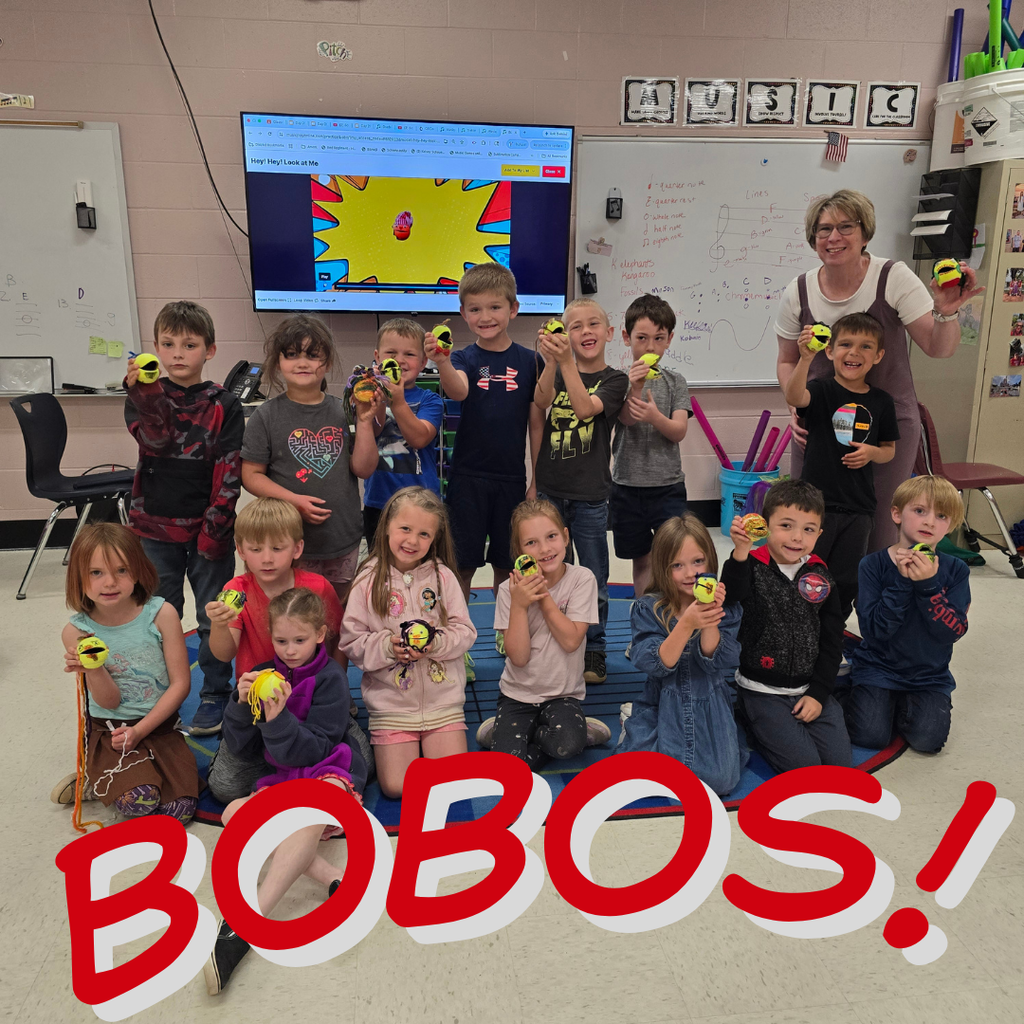 Children, some holding toys, sit and crouch in a classroom. A teacher stands behind them.
