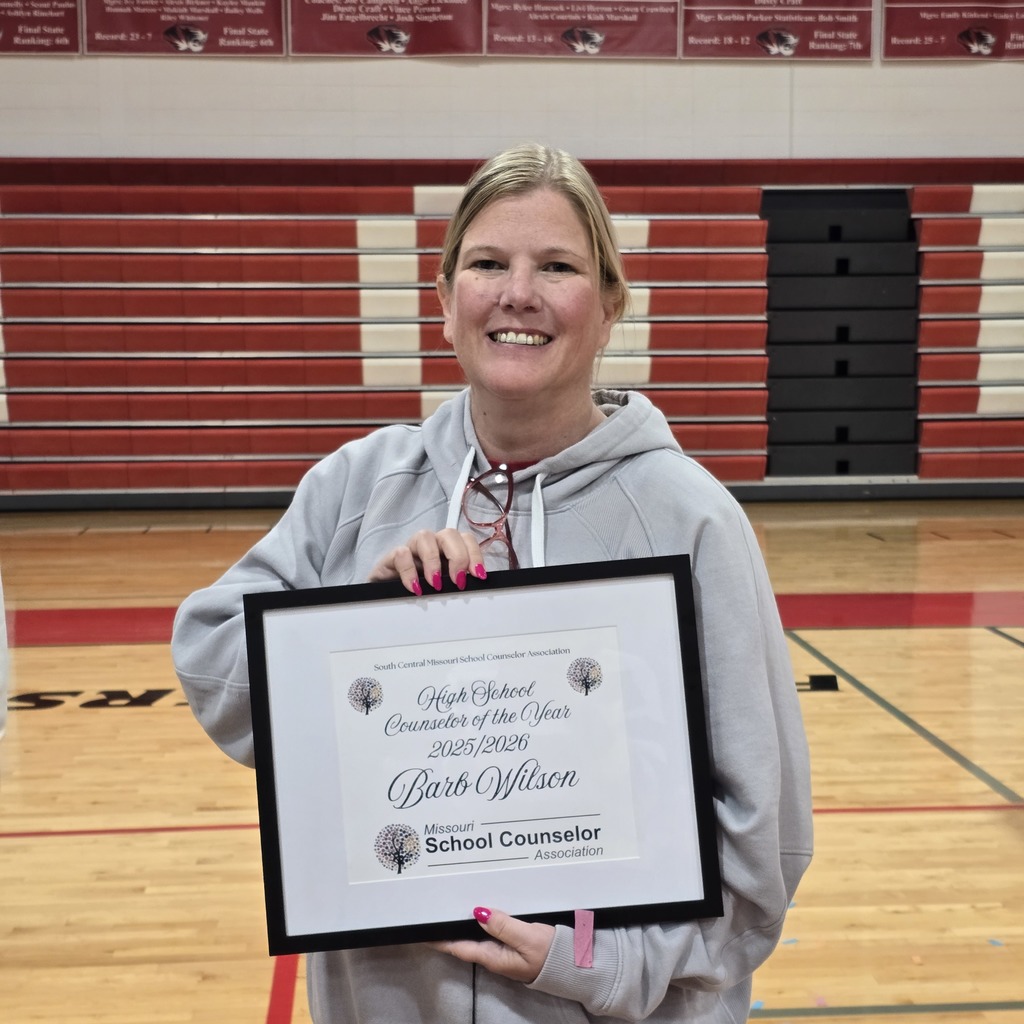 A woman holds a framed award certificate, smiling. She is wearing a gray hoodie. Behind her, a basketball court is visible.