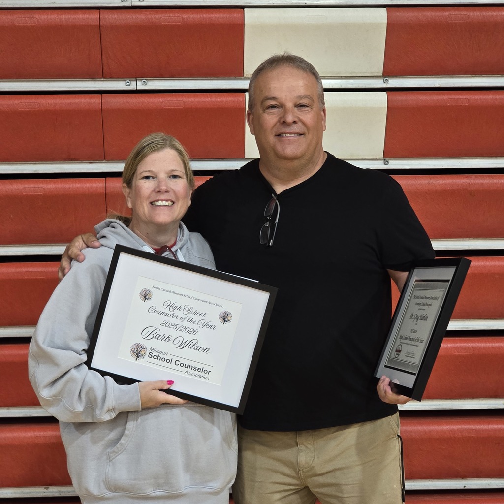 Two people, a woman and a man, standing in front of a red bleacher, each holding framed awards.