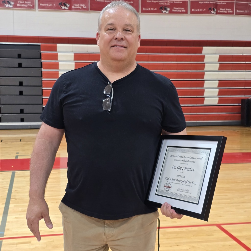 A man holds a framed certificate, wearing a black t-shirt and khaki pants, in a gym with a wooden floor and red and white bleachers.