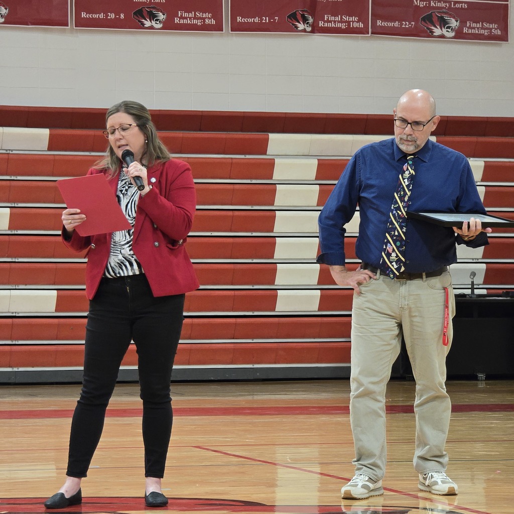 A woman in a red jacket and black pants stands on a basketball court, holding a red folder. An older man in a blue shirt and tie stands beside her.