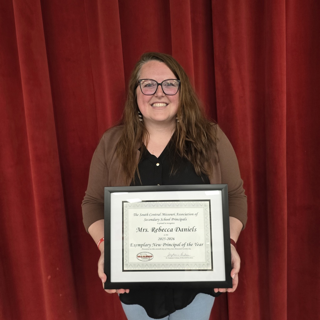 Woman with glasses holds a framed certificate in front of red theater curtains.