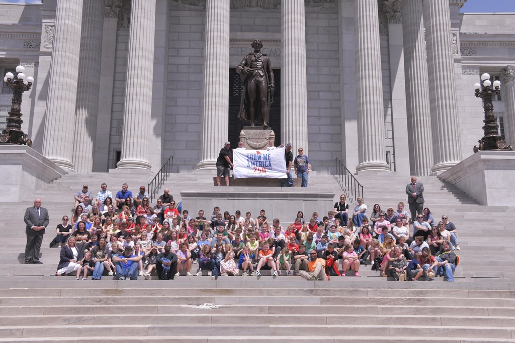 A large group of people is gathered on the steps of a building with columns, near a statue.
