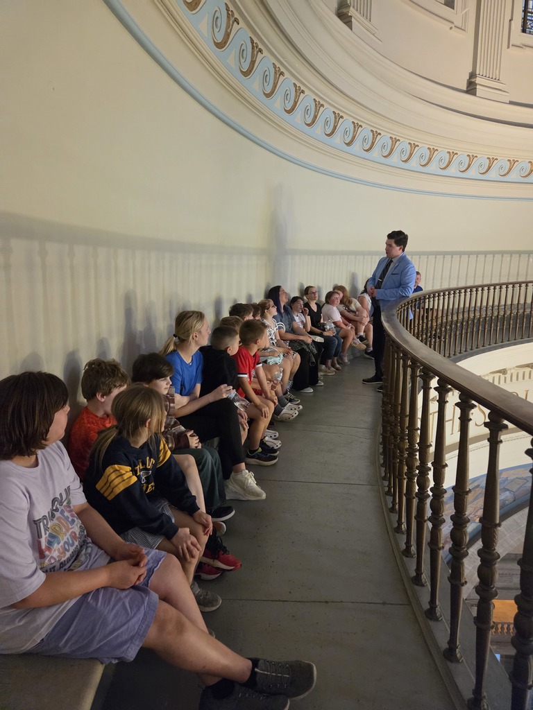 A group of children sit in a row on a bench while a man stands nearby. They are inside an ornate room.
