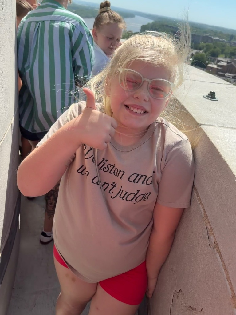 A girl in a beige t-shirt and red shorts smiles and gives a thumbs-up gesture on a balcony.