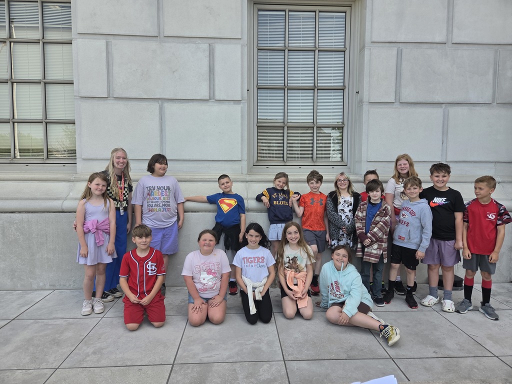 Group of children standing and sitting in front of a building with windows and a sidewalk.