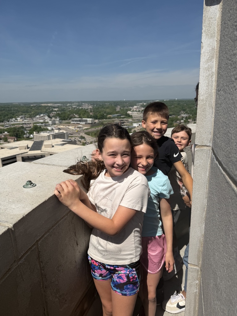 Five children standing on a building's ledge, with a city skyline visible behind them. One girl holds a dog.