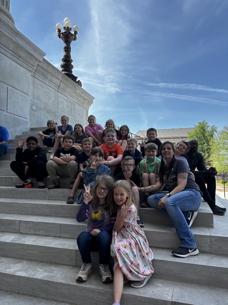 A group of children and adults pose for a photo on steps with a building and street lamp in the background.